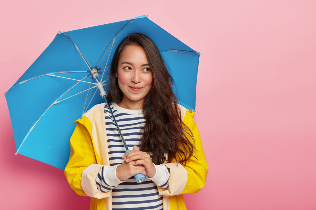 retty thoughtful woman with dark straight hair, holds blue umbrella, walks during cool day, protects herself from rain, wears yellow raincoat, enjoys September, poses over pink background